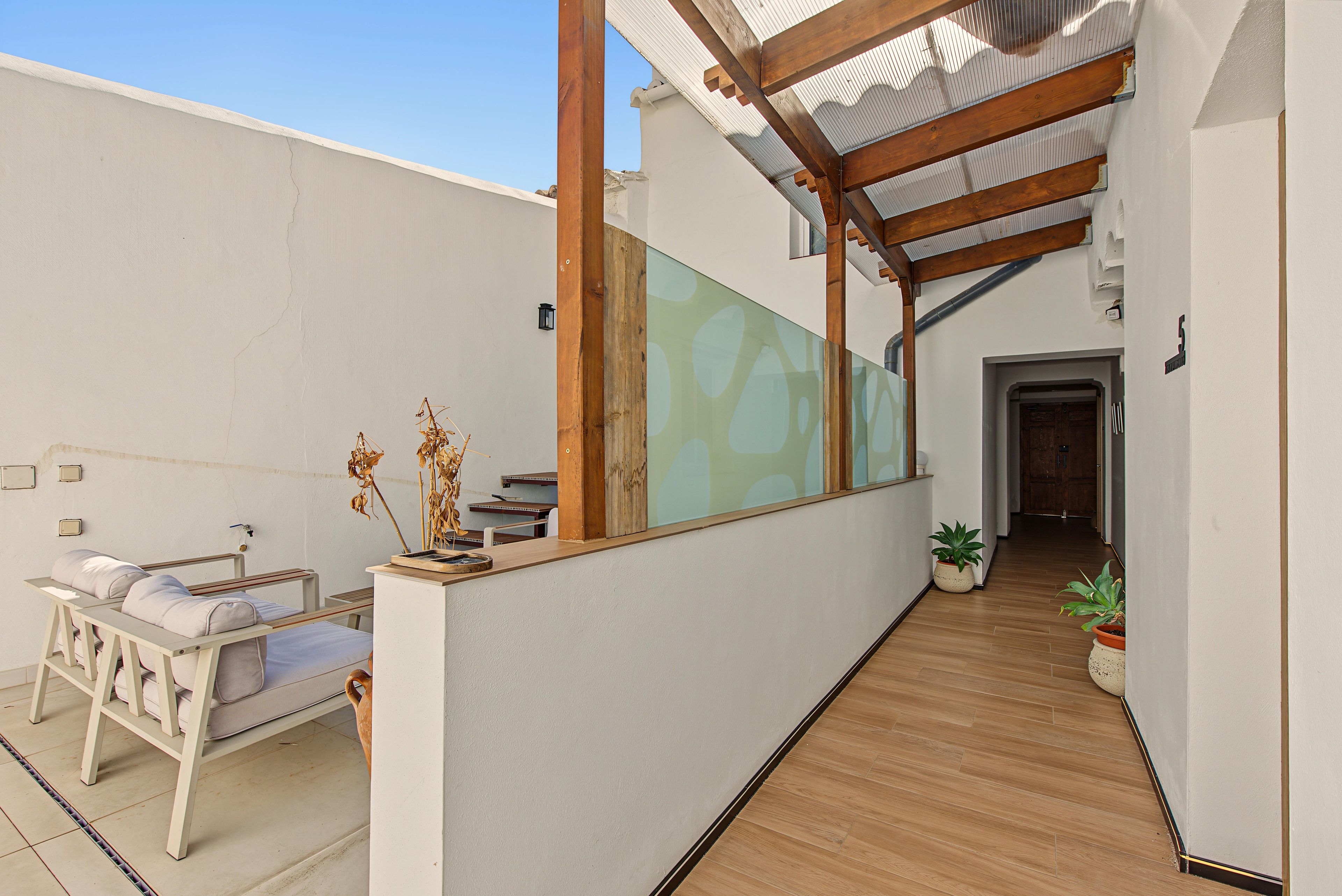 Un couloir doté d’un plancher en bois, d’un mur blanc et d’une cloison en verre. On y trouve un lit avec un matelas et des oreillers blancs, une petite table avec une lampe et une plante en pot.