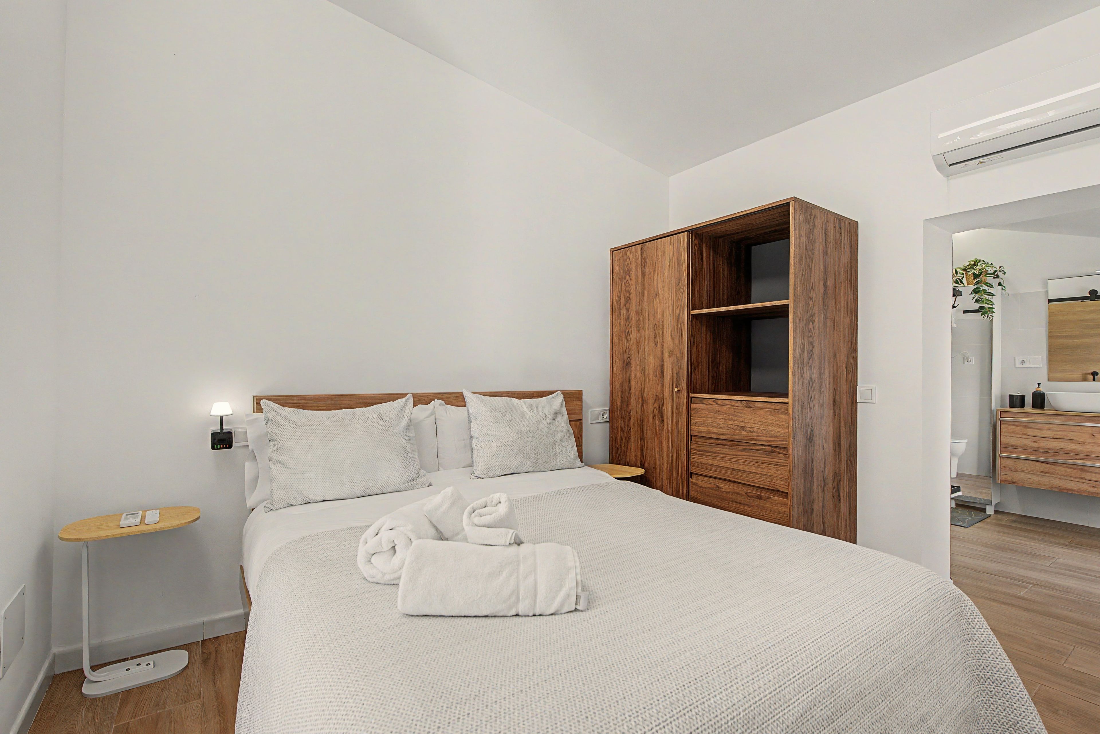 A hotel room with a bed, a wooden wardrobe, a bedside table, and a bathroom visible through an open door.