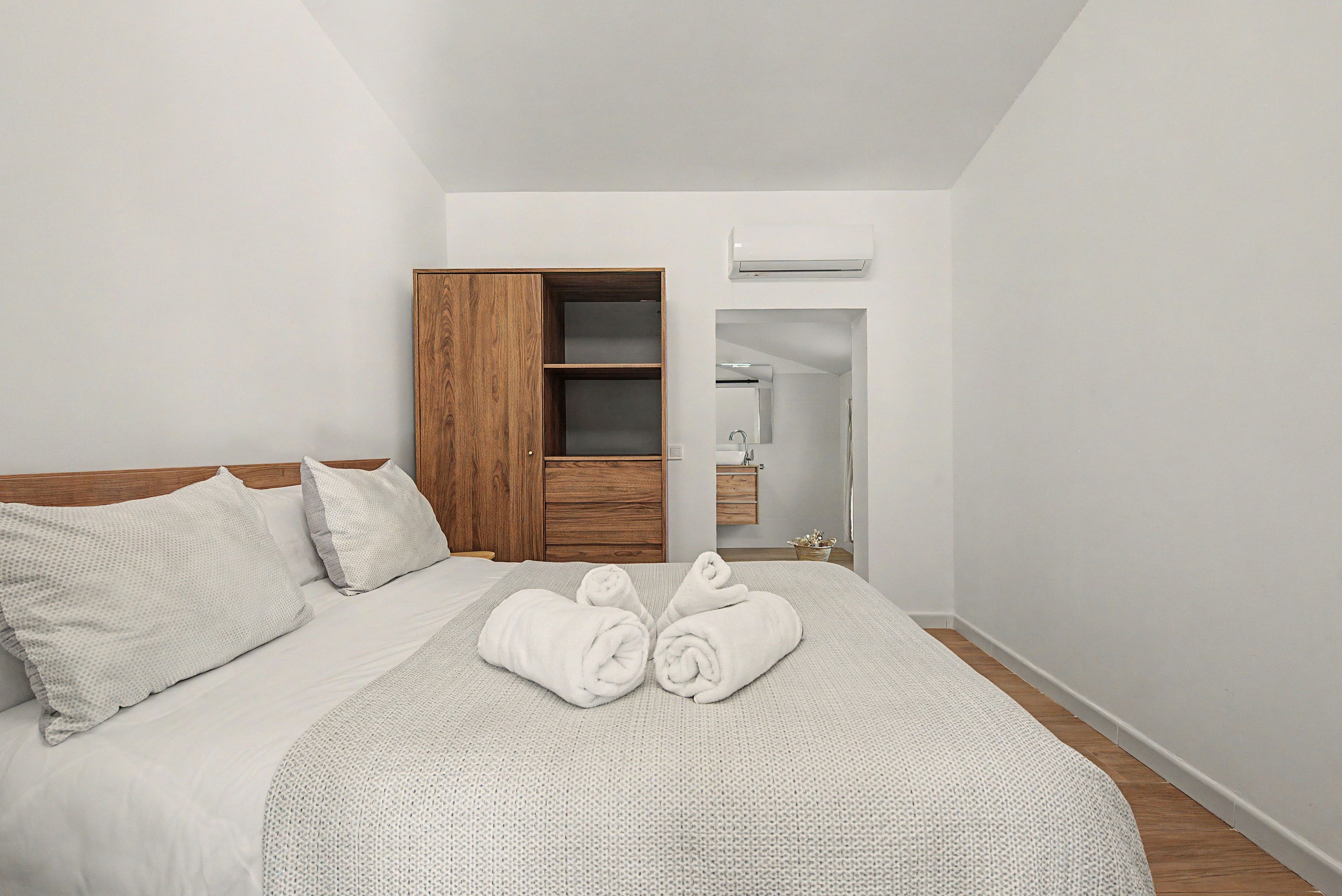 A bedroom with a bed, a wooden wardrobe, a bathroom, and a white air conditioning unit.