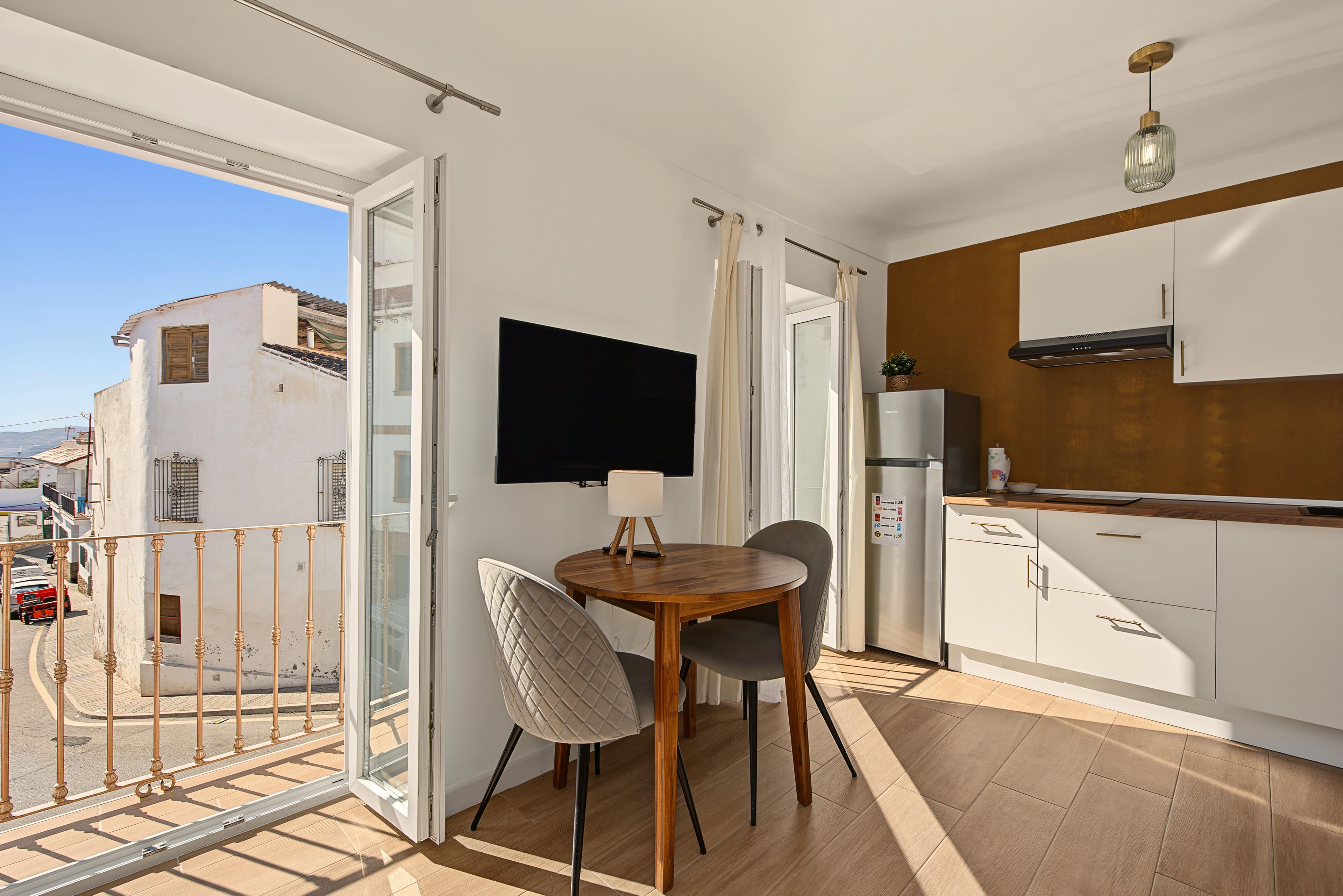 A balcony with a view of the street, a dining area with a table and chairs, a flat-screen TV mounted on the wall, and a kitchenette with cabinets and a stove.