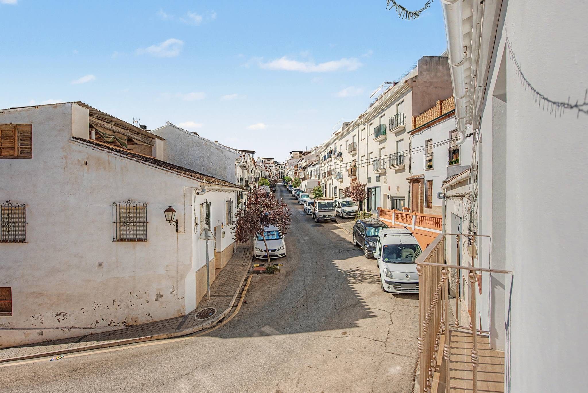 Eine enge Straße, gesäumt von weißen Gebäuden und geparkten Autos, unter blauem Himmel.