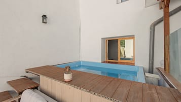 Een houten terras met een zwembad, een kleine tafel met een bloempot en een raam met uitzicht op groen.