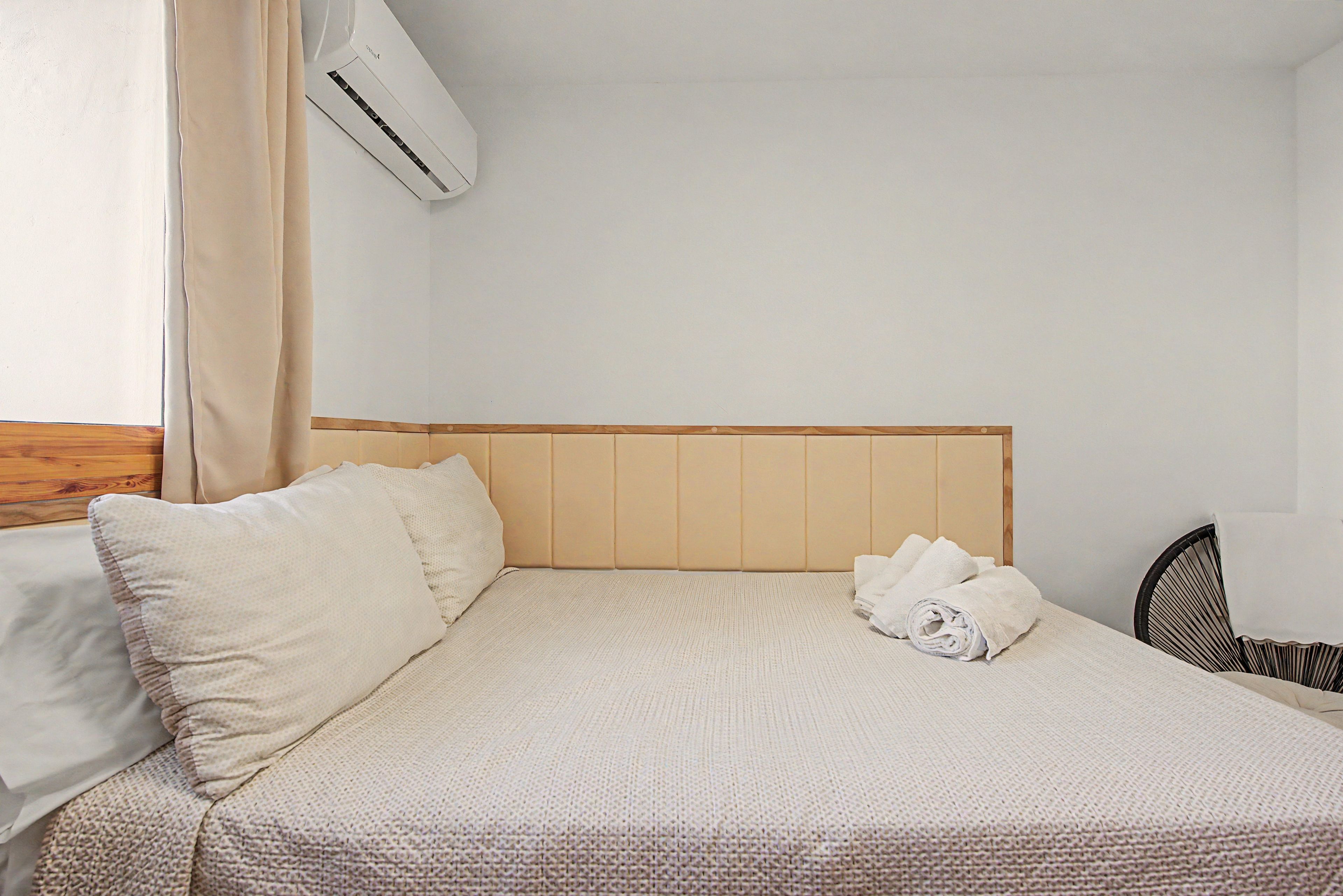 Ein ordentlich bezogenes Bett mit Kissen und einem zusammengerollten Handtuch, ein Holz-Kopfteil und ein Fenster mit Vorhängen.