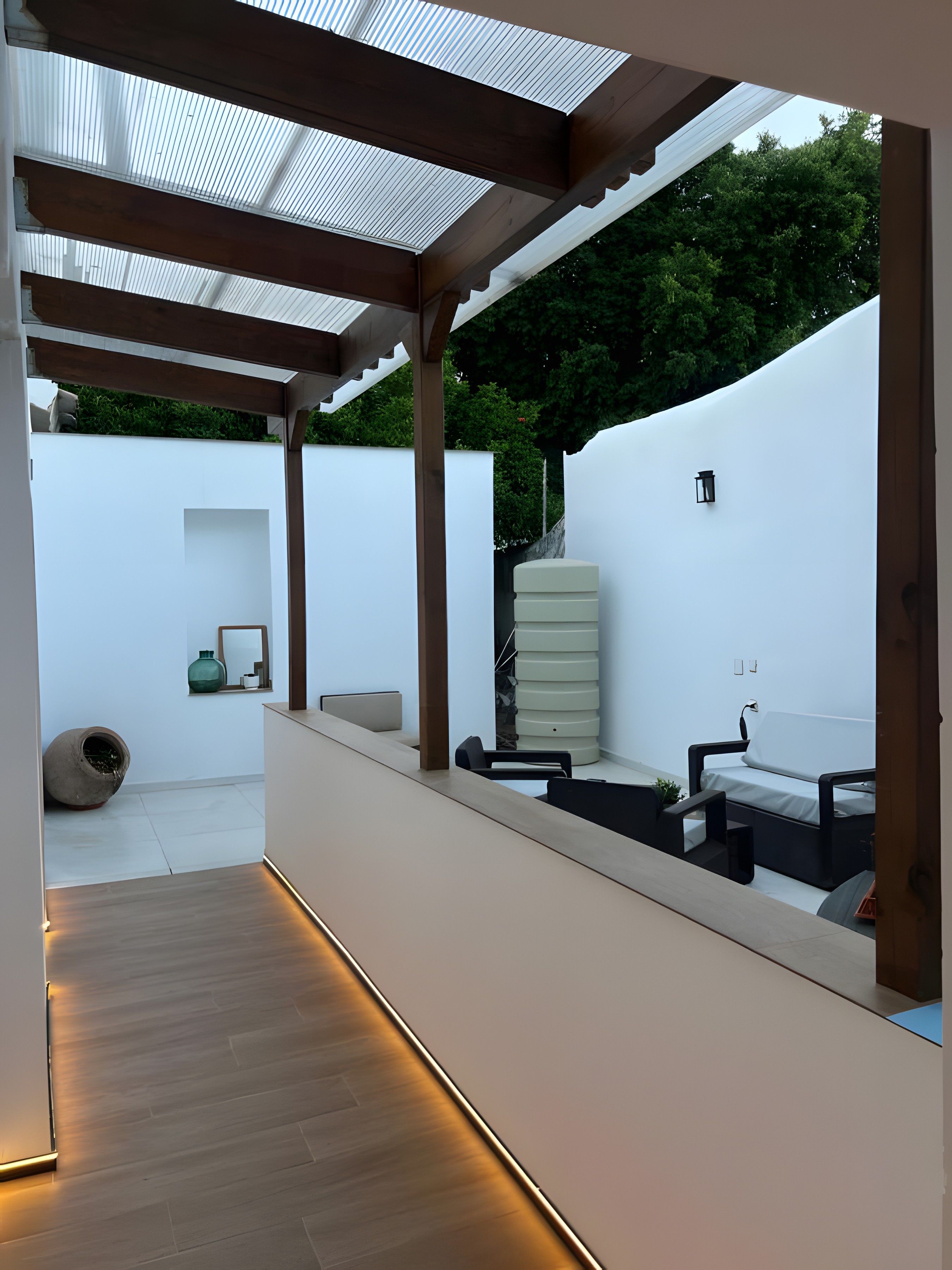 Un patio moderno al aire libre con una pérgola de madera, pared blanca y una zona de estar con sofá blanco y sillas negras.
