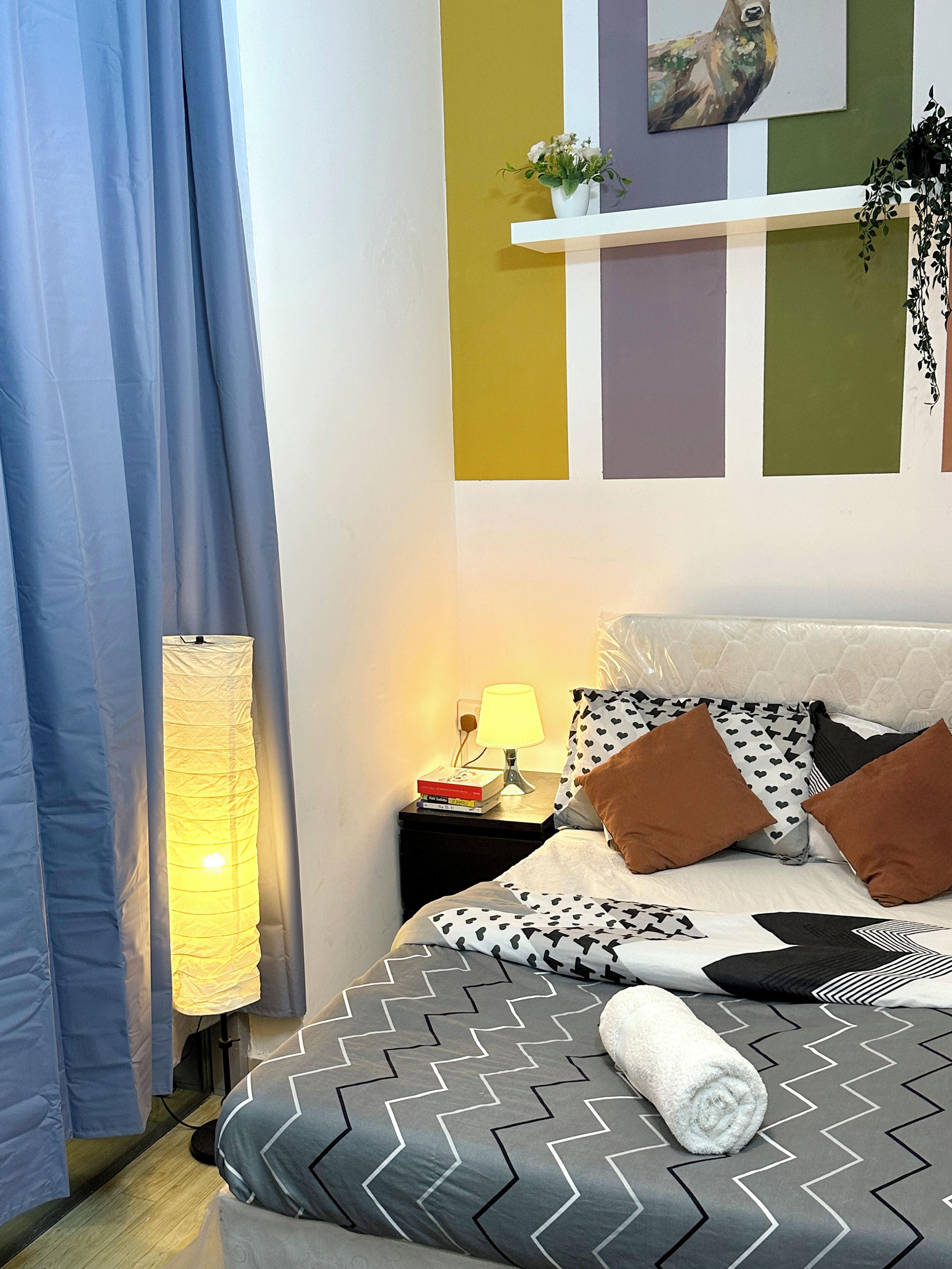Een keurig opgemaakte slaapkamer met een bed, kussens, een bedlampje, een nachtkastje en een muur met een schilderij en een plank met een plant.