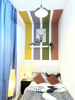 A bedroom with a bed, a nightstand, a lamp, a wall-mounted shelf with a plant, and a painting of a deer. - Twin Bed Partition Room Near ICM Metro (Dubai)