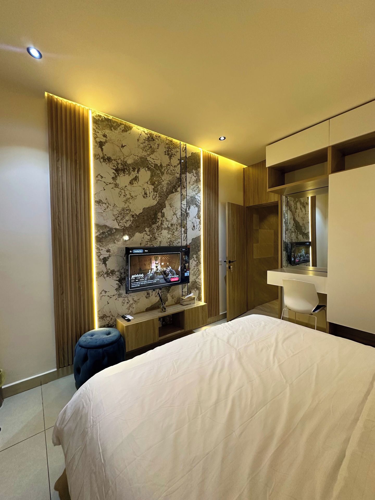 A modern hotel room with a large bed, a TV mounted on a marble wall, a desk with a chair, and a wardrobe.