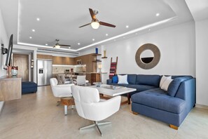 Een moderne woonkamer met een blauwe bank, witte stoelen, een glazen salontafel en een keuken op de achtergrond.