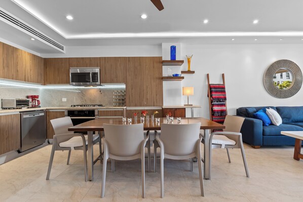 Een moderne keuken met inbouwapparatuur, een eethoek met tafel en stoelen, en een woonkamer met een bank en een decoratieve spiegel.