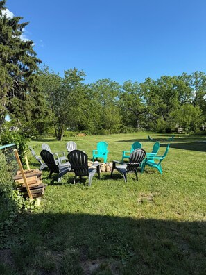 Ein Sitzbereich im Freien mit Adirondack-Stühlen und einem Holztisch, umgeben von Bäumen und blauem Himmel.