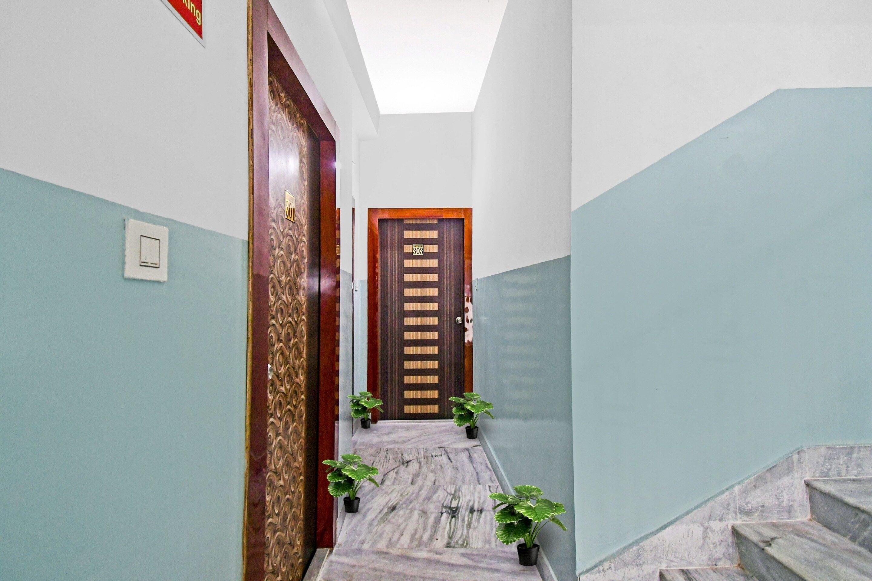 Un pasillo con piso de mármol, una puerta de madera con placa de número y dos plantas en macetas.