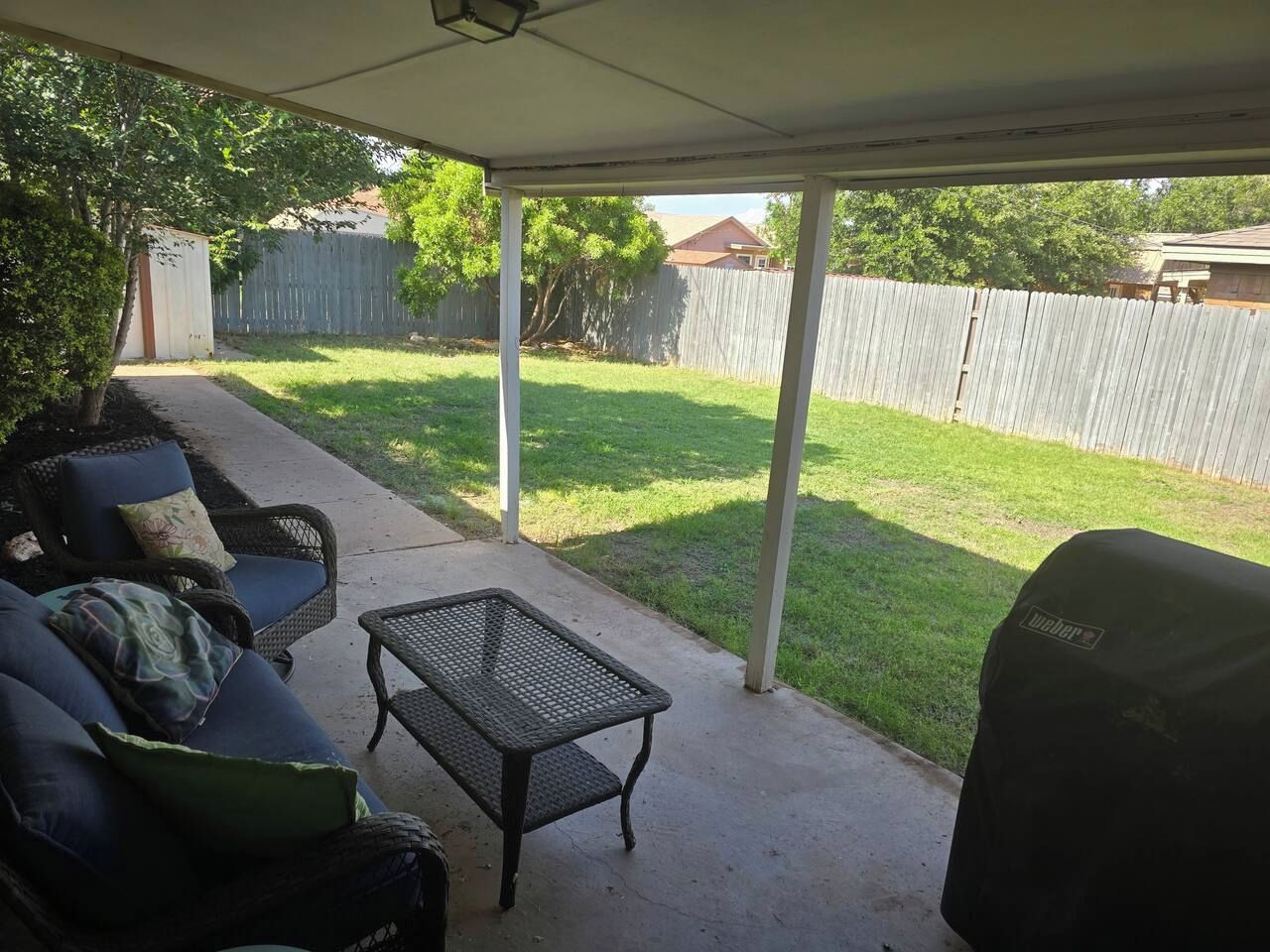 Een overdekt terras met een barbecue, tuinmeubilair en uitzicht op een achtertuin met een houten schutting en gras.