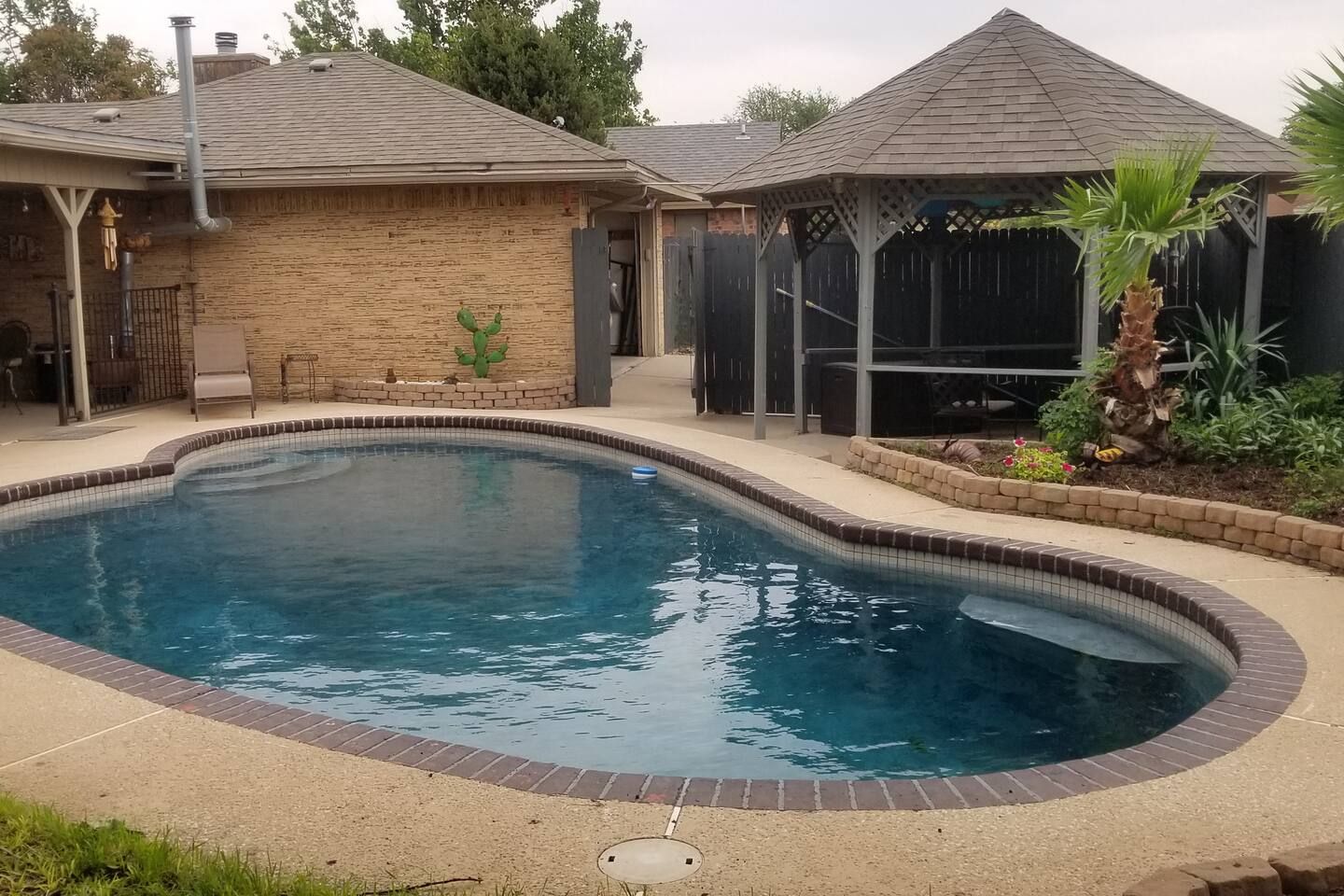 A residential swimming pool with a curved design, surrounded by a brick deck and a covered patio area.