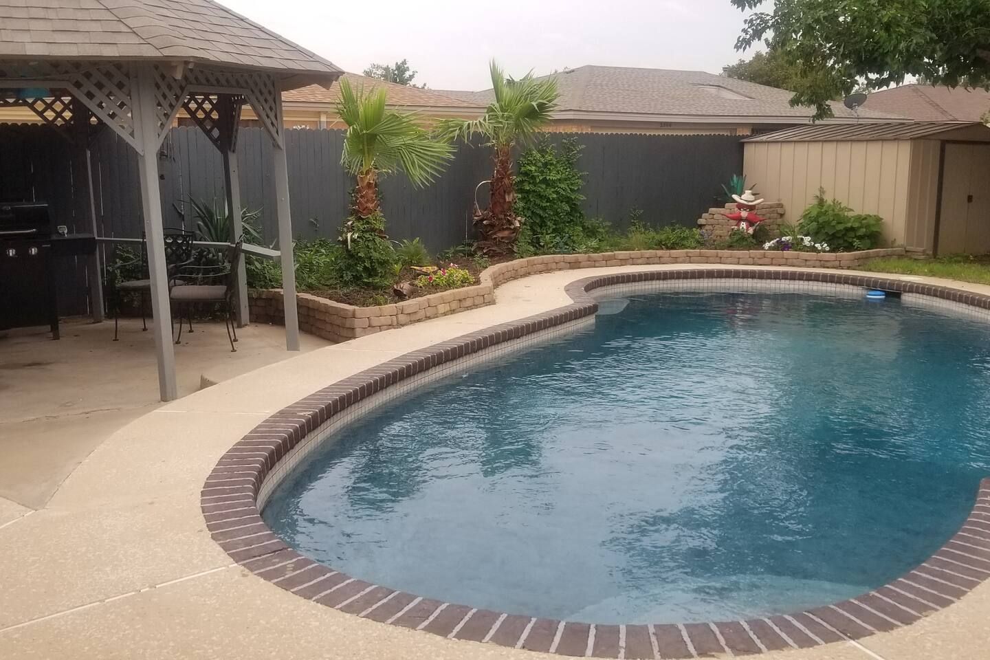 Una piscina con borde de ladrillo, rodeada por un patio con pérgola y mobiliario exterior.