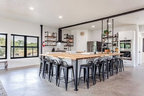 A modern kitchen with a central island, bar stools, and a view through large windows. - Xenia Ranch Barndo l Pool HotTub l Partial House (Valley Mills)