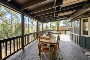 A covered outdoor deck with a table and chairs, surrounded by a railing, overlooking a forested area. - Gonzo Chalet (Ruidoso)