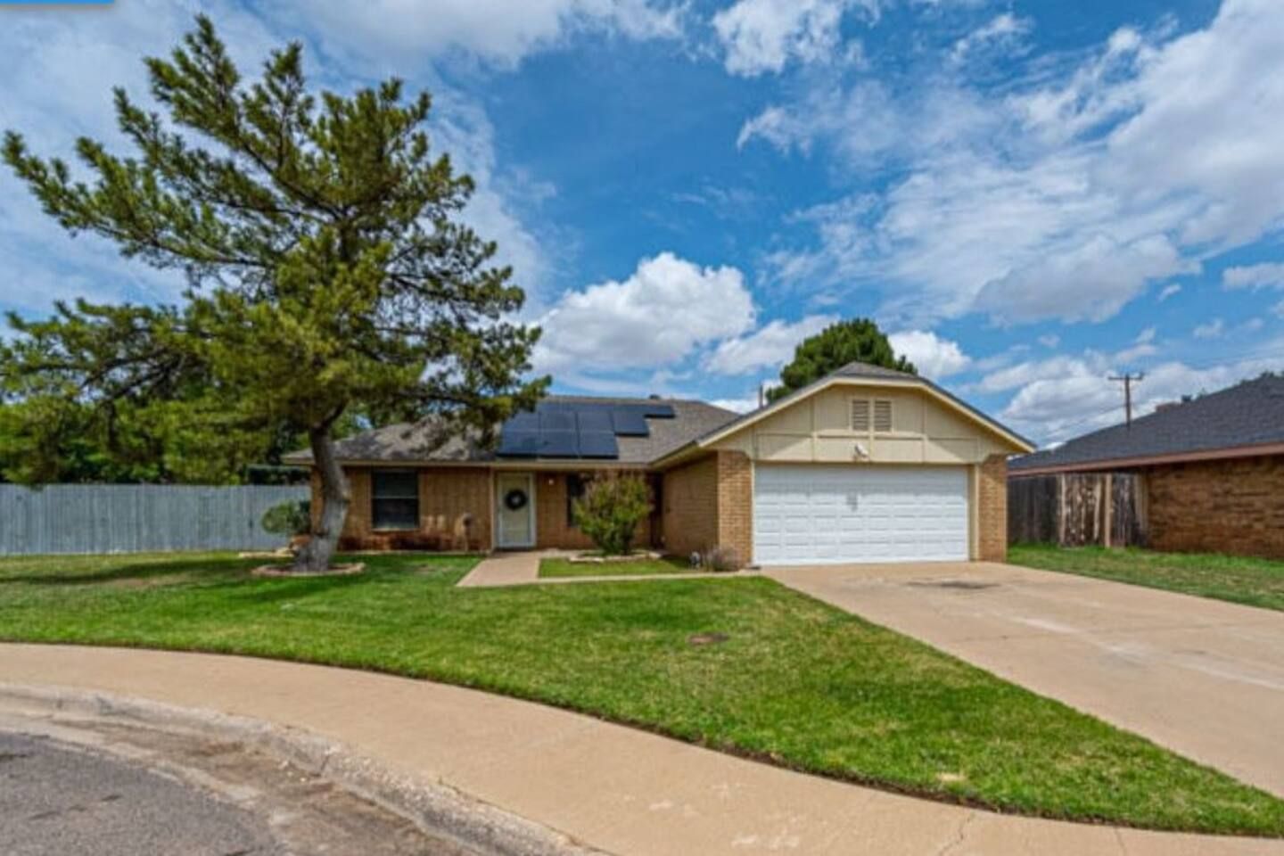A single-story house with a garage, solar panels on the roof, a well-maintained lawn, and a large tree in front.