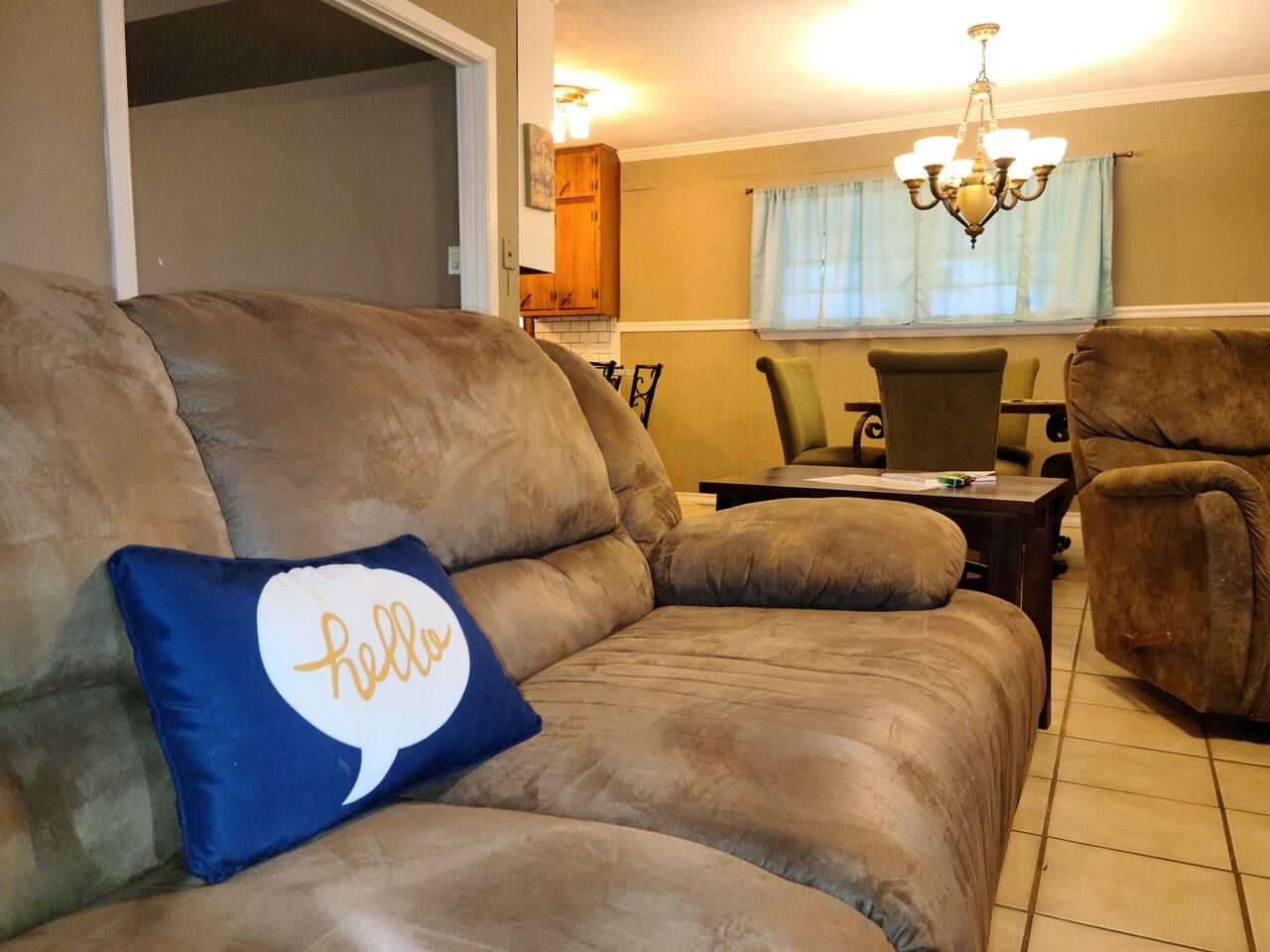 A living room with a brown sofa, a blue pillow with 'hello' text, a dining table, chairs, and a chandelier.