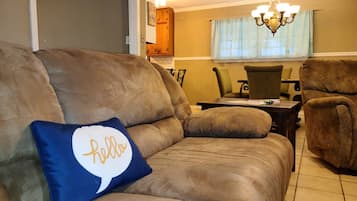 A living room with a brown sofa, a blue pillow with 'hello' text, a dining table, chairs, and a chandelier.