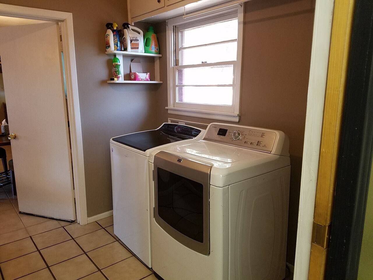 A laundry room with a front-loading washer and dryer, a window above the dryer, and a shelf with cleaning supplies.