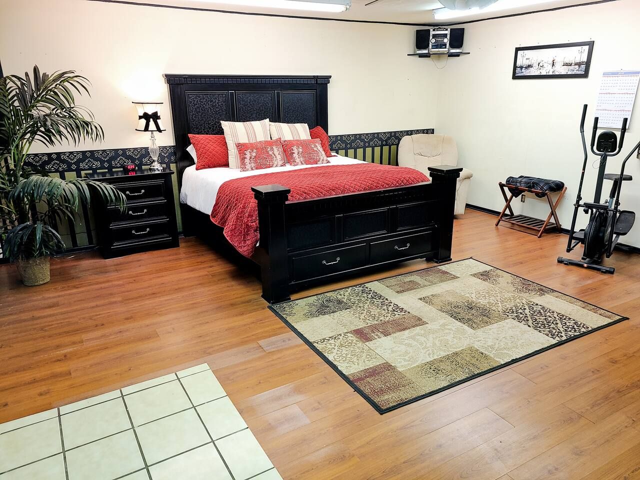 Una camera da letto con un letto, un comodino, una lampada, una sedia, un tavolino, una pianta, un attrezzo per esercizi e un tappeto.