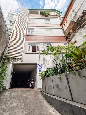 Une ruelle étroite menant à l’entrée d’un immeuble, avec une porte de garage, le tout bordé de végétation et entouré d’un mur en béton.