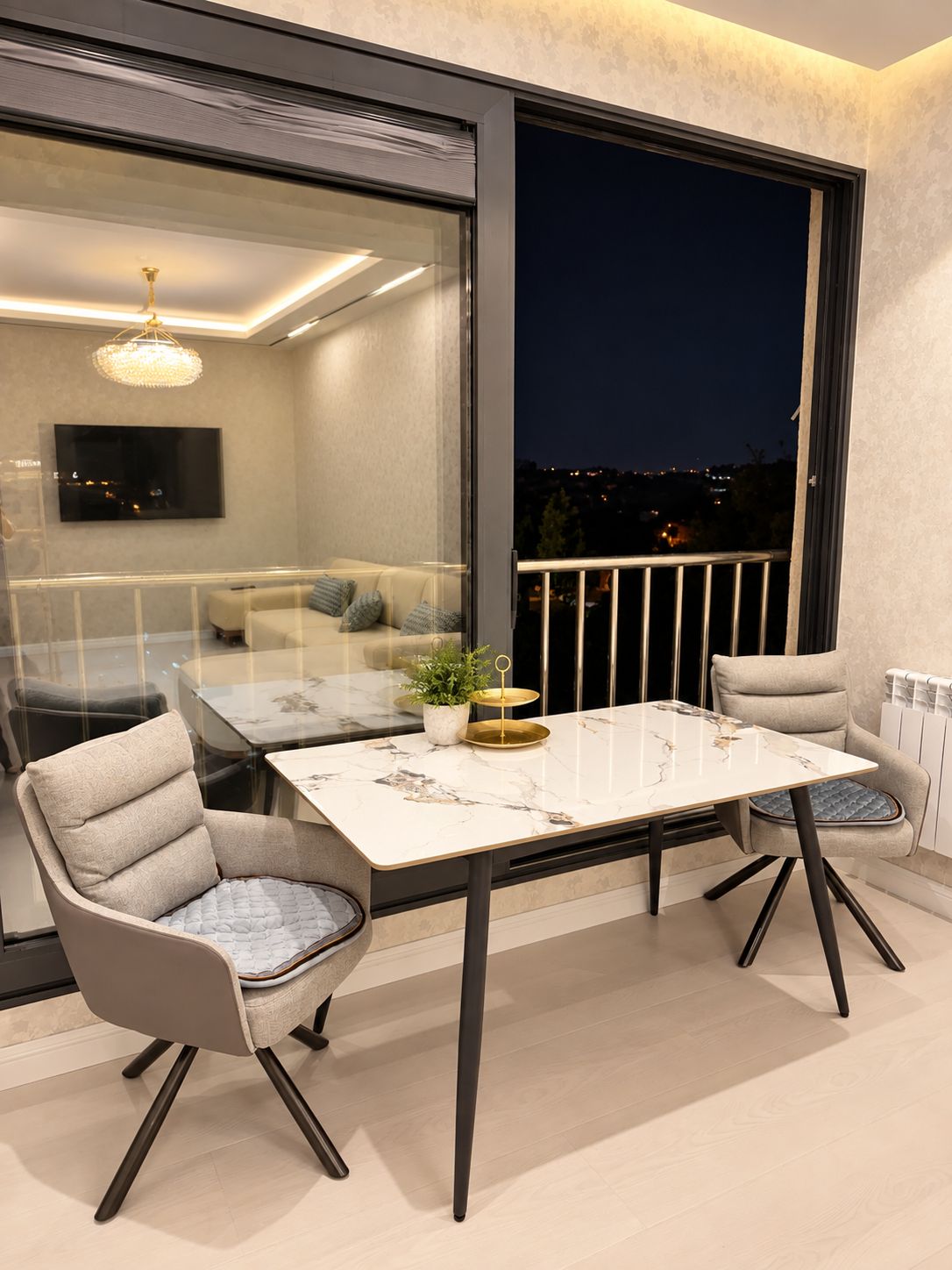 A balcony with a glass table and chairs, a potted plant, and a view of the city at night.