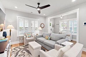 A modern living room with a grey sofa, white armchairs, a ceiling fan, and large windows with shutters. - Coastal Charm in Downtown Wilmington 2BR, Walk to Dining & Riverfront (Wilmington)