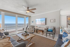 A spacious living room with a large window overlooking the beach, a ceiling fan, and a flat-screen TV mounted on the wall. - Smyrna Beach Club 218b (New Smyrna Beach)