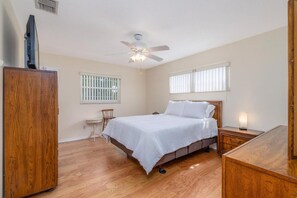 A bedroom with a bed, a ceiling fan, a nightstand, a wooden wardrobe, and a small table with a chair. - Pet friendly home with large backyard (New Smyrna Beach)