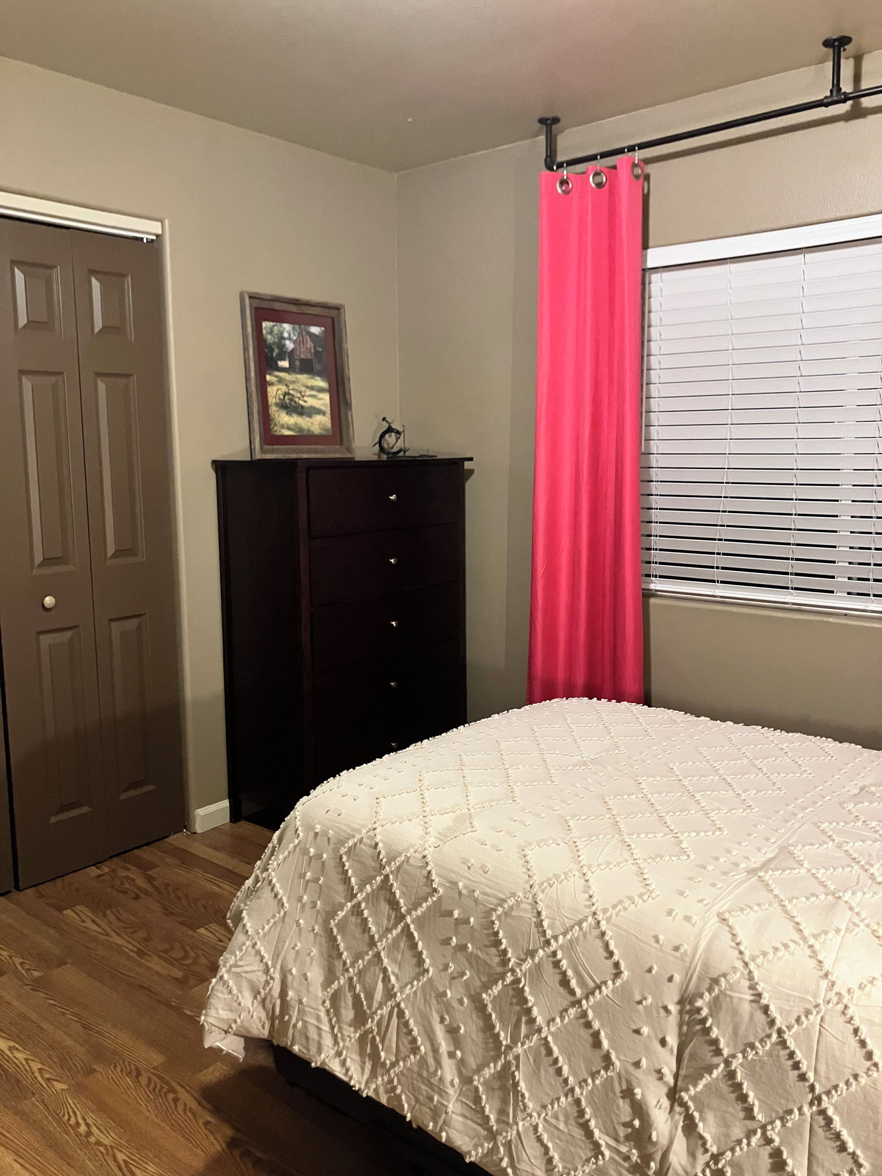 A bedroom with a bed, a dresser, a door, and a window with blinds.