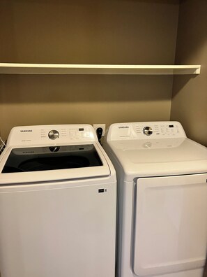 Two white Samsung front-loading washing machines in a laundry room with a beige wall and a white shelf above.