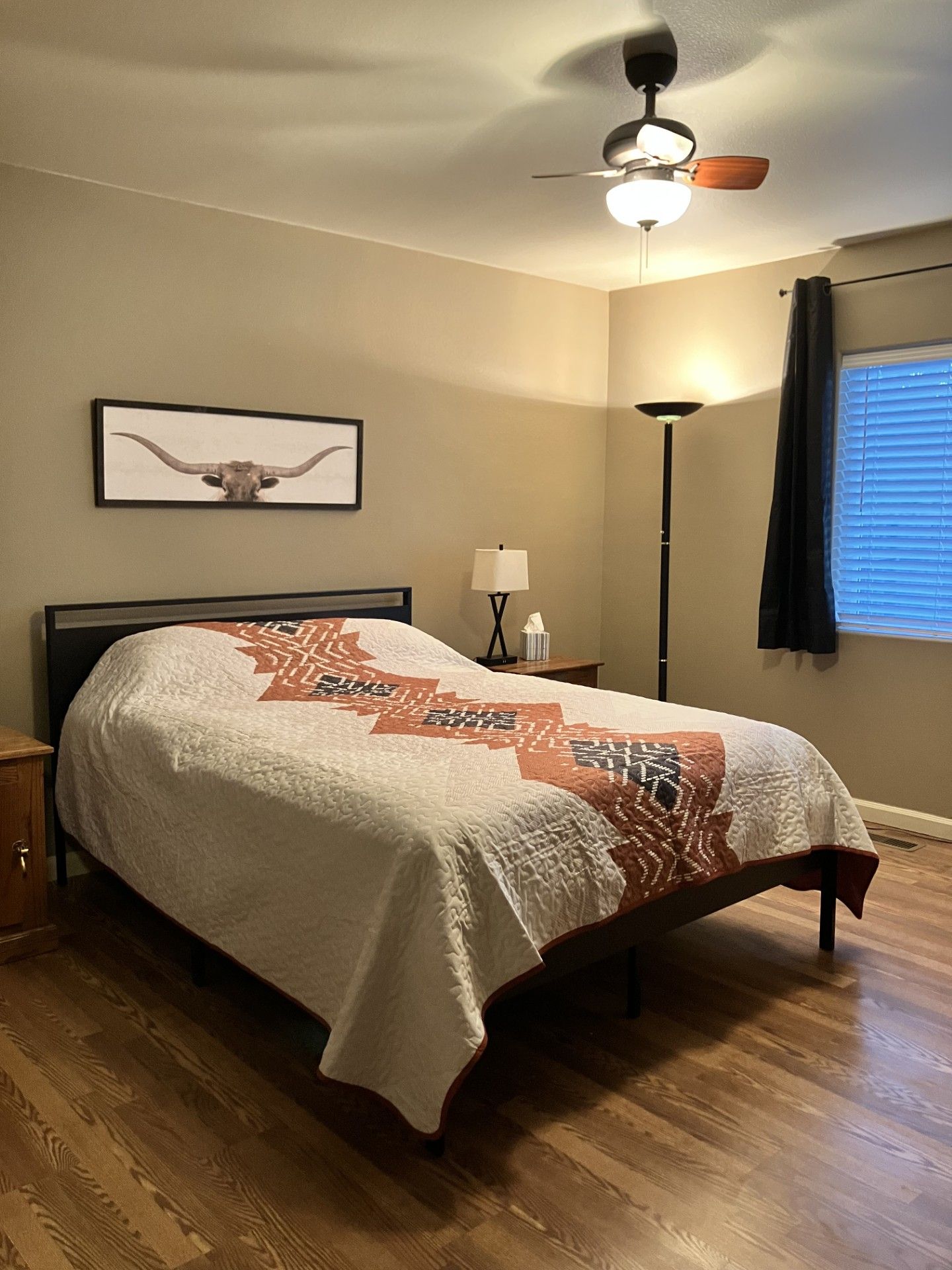 Een slaapkamer met een bed, een plafondventilator, een bedlampje, een ingelijst schilderij aan de muur, en een raam met zonwering.