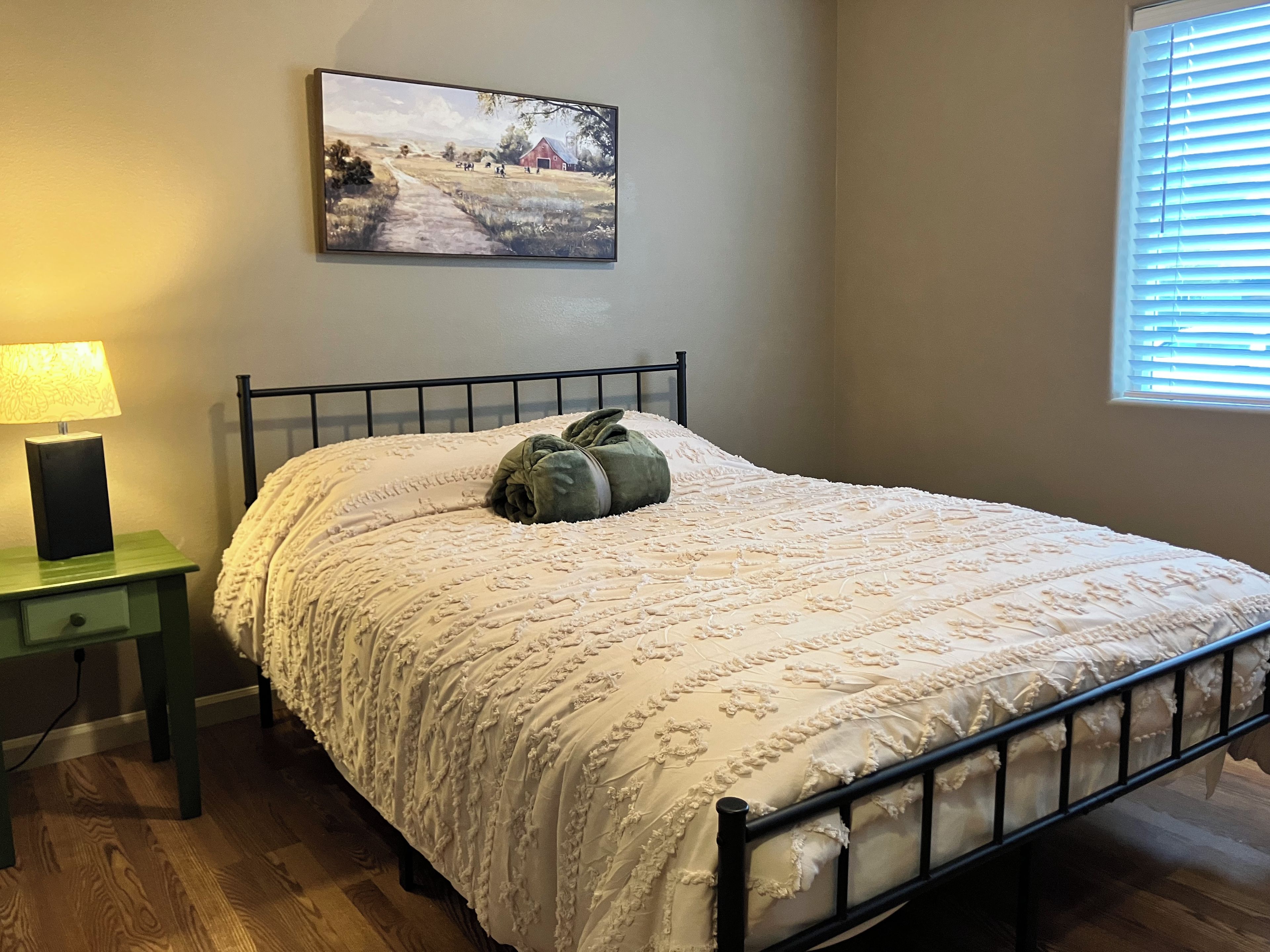 Une chambre à coucher avec un lit, une table de chevet, une lampe, un tableau au mur et une fenêtre avec des stores.