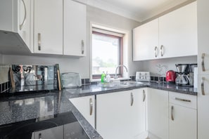 A modern kitchen with white cabinets, a granite countertop, and stainless steel appliances including a coffee maker and toaster. - Charming 3-Bed Home with Garden and Parking (Cambridge)