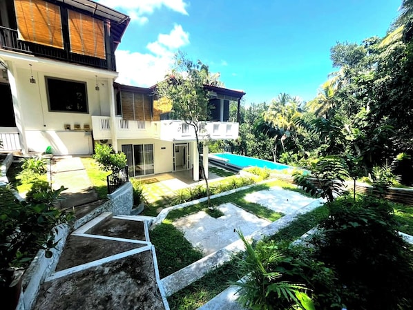 Una casa moderna de dos pisos con piscina, rodeada de exuberante vegetación y un jardín cuidado.