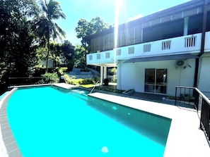 Ein Pool mit klar blauem Wasser, umgeben von einem weißen Gebäude mit Balkon und Palmen im Hintergrund.