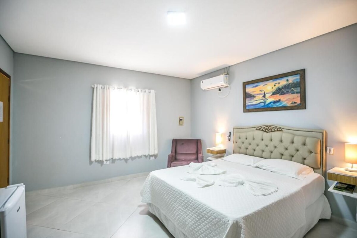 A hotel room with a bed, a chair, a painting, an air conditioner, and a window with curtains.