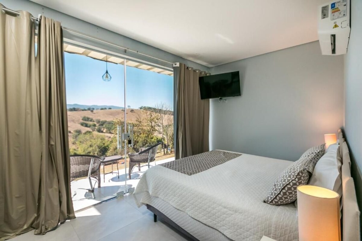 A bedroom with a bed, a TV, a balcony with a chair, and a view of the countryside.