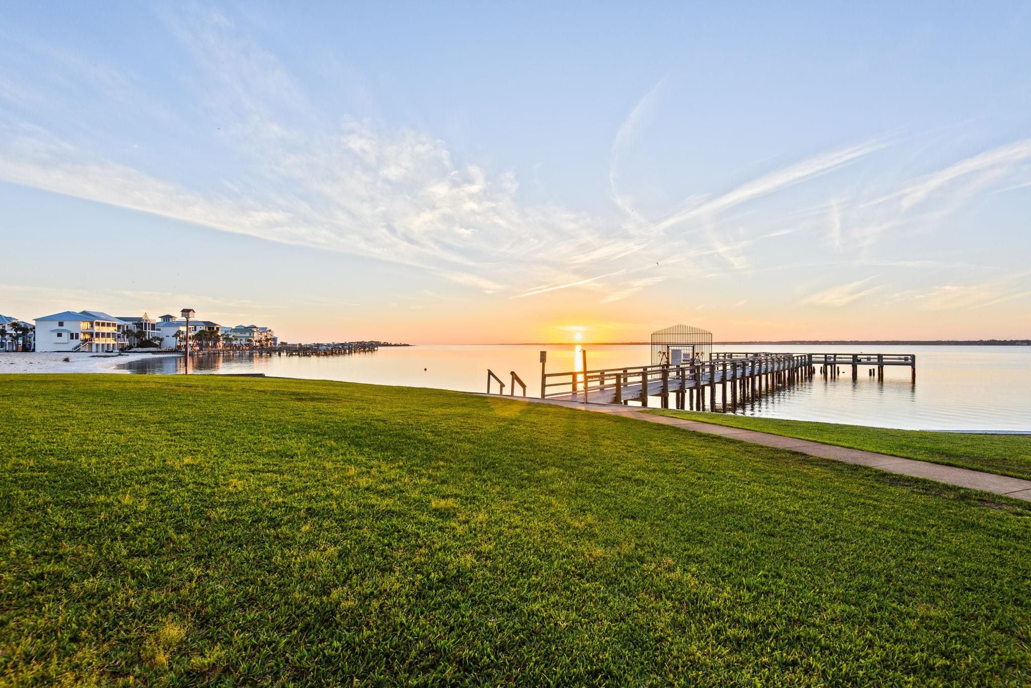 A grassy area with a wooden pier extending into calm water, a clear sky, and buildings in the background.
