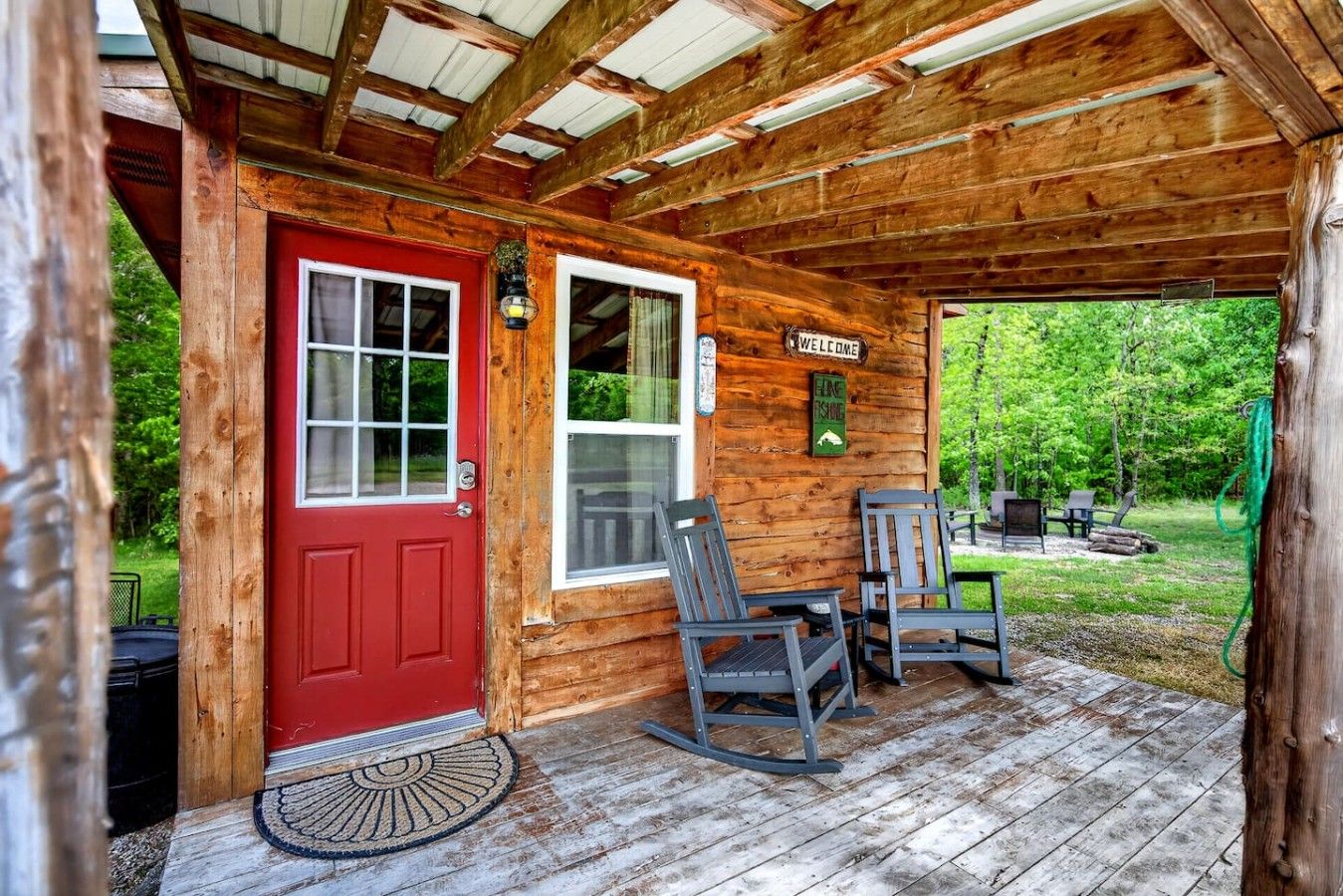 A wooden cabin with a red door, two rocking chairs, a welcome sign, and a covered porch.
