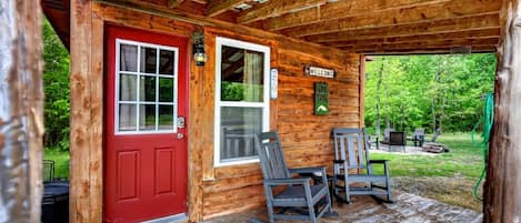 A wooden cabin with a red door, two rocking chairs, a welcome sign, and a covered porch.