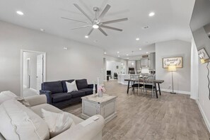 A modern living room with a dark sofa, a white armchair, and a coffee table. There is a dining area with a table and chairs, and a kitchen in the background. - Riverfront Denton Retreat | Spacious 4BR (Denton)