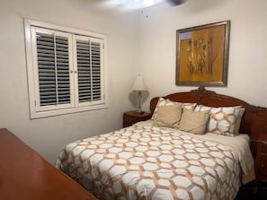 Un dormitorio con cama, mesita de noche, lámpara, ventana con contraventanas y un cuadro en la pared.