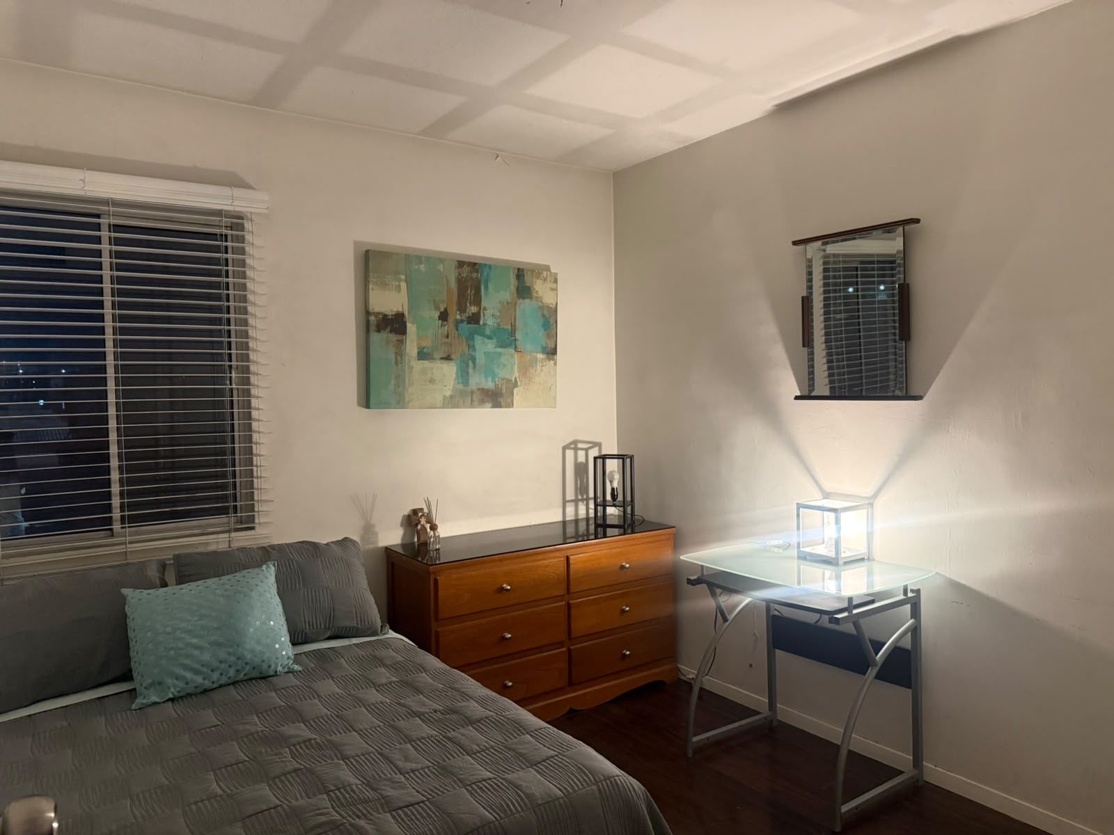 A bedroom with a bed, dresser, desk, and artwork on the wall.