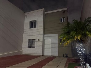 Un edificio de dos plantas con fachada blanca, un primer piso rojo y color beige, y un pequeño jardín con una palmera y luces colgantes.