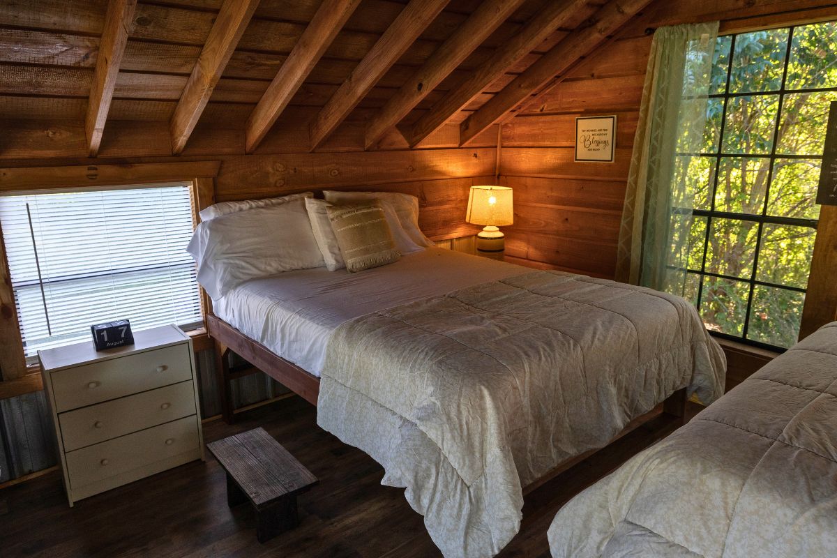 A wooden cabin bedroom with a bed, bedside table, lamp, and window with curtains.