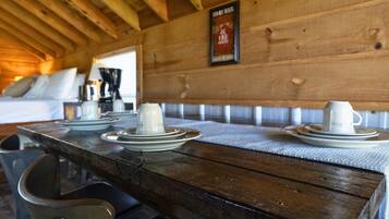 Une table de salle à manger en bois, avec des assiettes et des tasses disposées pour un repas, un banc et un lit en arrière-plan.