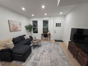 Un salon moderne avec un canapé, une table basse et un téléviseur. On y trouve un bureau avec une chaise et une plante. Les murs de la pièce sont blancs et il y a une grande fenêtre.