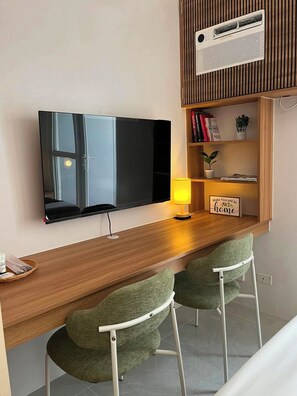 A modern living space with a flat-screen TV mounted on the wall, a wooden desk with a lamp, two green upholstered chairs, a bookshelf with books and a small plant, and a sign that reads 'Make Yourself At home'. - Ground Floor Studio with Patio and Pool View at Casa Mira Towers, Mandaue (Mandaue)